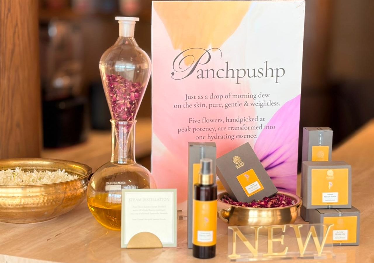 Panchpushp steam distillation table with a glass still, rose petals, brass urlis and the NEW sign